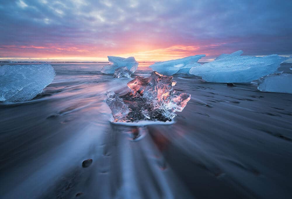 Large clear iceberg in centre of image, surrounded with white icebergs against pink and orange sunset backdrop
